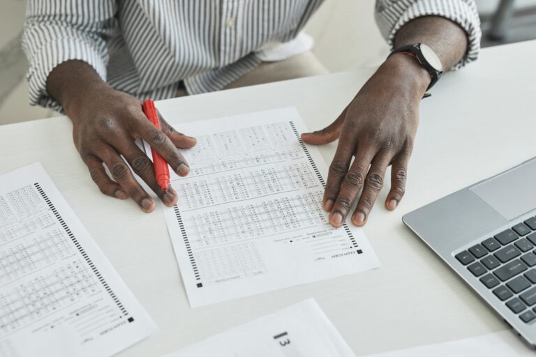 An educator carefully checks exam papers using a red marker at a desk, illustrating focused grading.